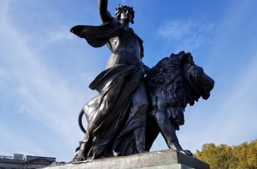 A bronze statue of a woman walking by a lion holding an olive branch aloft. This is the "Peace" sculpture, one of four large bronze corner groups belonging to the Victoria Memorial immediately outside Buckingham Palace.
