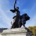 A bronze statue of a woman walking by a lion holding an olive branch aloft. This is the "Peace" sculpture, one of four large bronze corner groups belonging to the Victoria Memorial immediately outside Buckingham Palace.