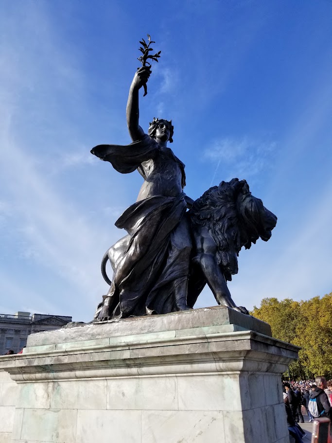 A bronze statue of a woman walking by a lion holding an olive branch aloft. This is the "Peace" sculpture, one of four large bronze corner groups belonging to the Victoria Memorial immediately outside Buckingham Palace.