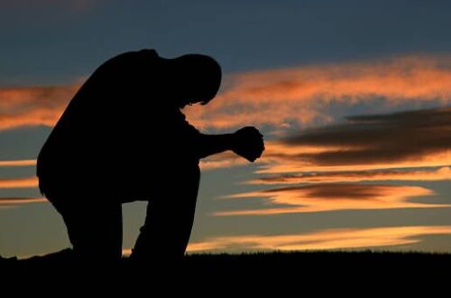 Photo of a man silhouetted on one knee, hands clasped in reverent prayer, with the sun just beginning to rise in the background.