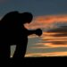 Photo of a man silhouetted on one knee, hands clasped in reverent prayer, with the sun just beginning to rise in the background.