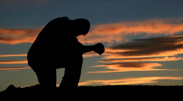 Photo of a man silhouetted on one knee, hands clasped in reverent prayer, with the sun just beginning to rise in the background.