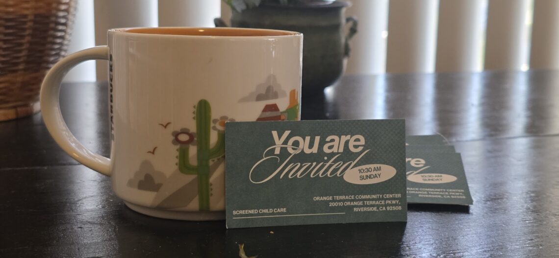 A close-up of a church card invitation propped up against a coffee cup on a black table with plants in the background.