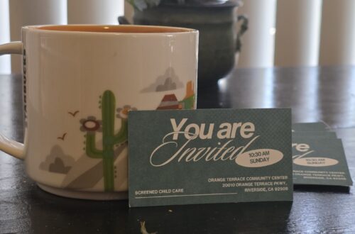 A close-up of a church card invitation propped up against a coffee cup on a black table with plants in the background.